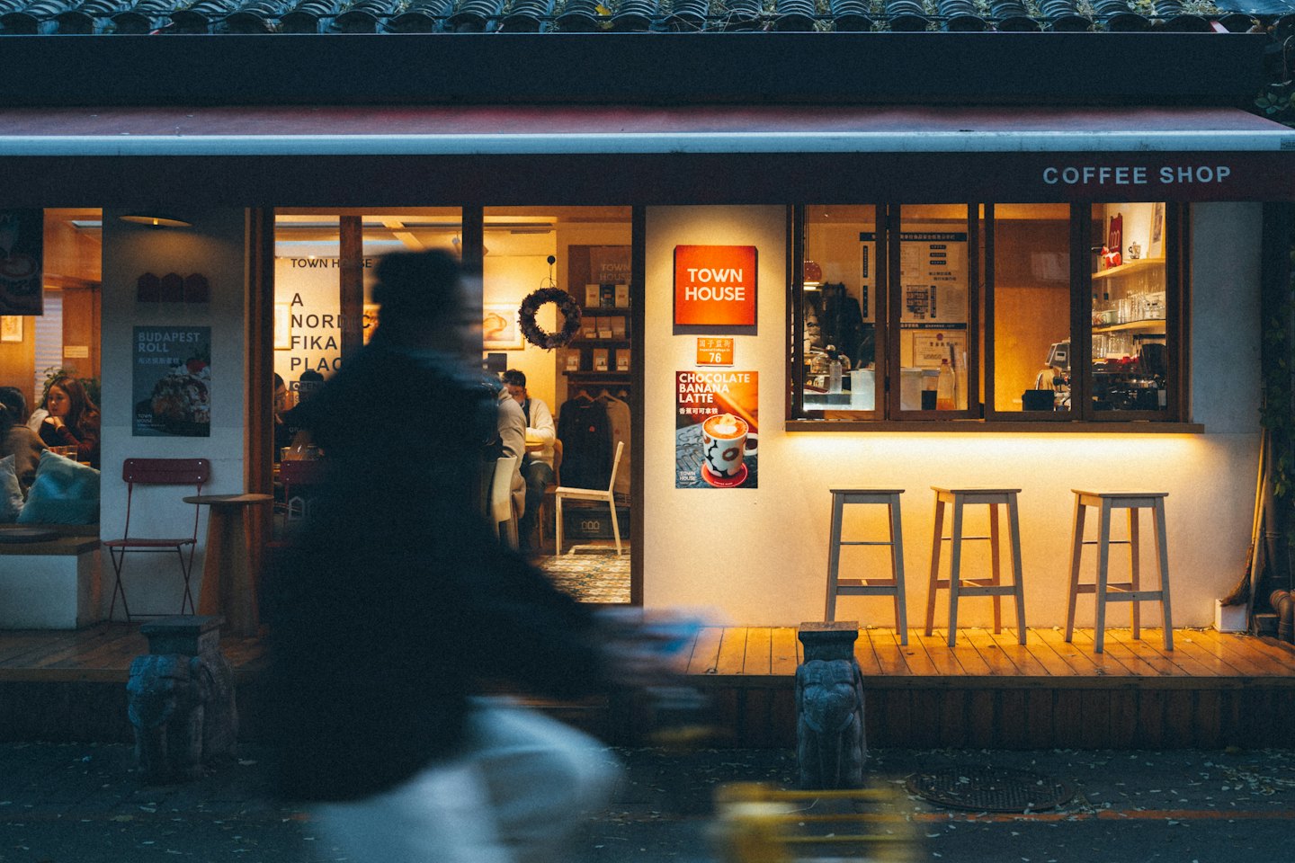 Coffee shop with red awning at dusk, customers inside.